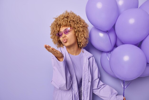 Fashionable curly haired woman blows air kiss expresses love\
wears suglasses and jacket holds bunch of inflated balloons\
celebrates special occasion isolated over purple background empty\
space