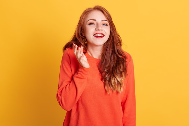 Fashionable European happy woman with wavy hair, wearing orange sweater, raises palms, posing on yellow wall