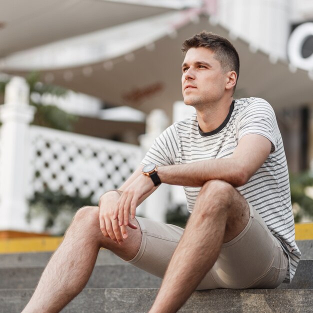 Photo fashionable european young man in a stylish striped t-shirt in summer beige shorts sits on the stairs on the street near a vintage outdoor cafe.