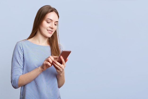 Fashionable female in stylish clothes, holds smart phone, checks her bank account, poses against blue wall with copy space for your promotional text or advertisement. Technology and people