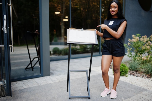 Fashionable feminist african american woman wear in black t-shirt and shorts, posed outdoor and show menu of restaurant.