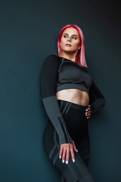 Fashionable fitness trainer model woman with pink hair in black sportswear stands near a black wall