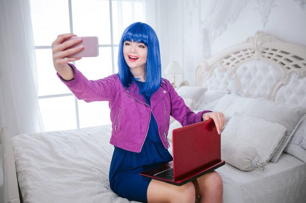 Fashionable freak. Glamour smiling beautiful woman with blue hair and trendy makeup is holding computer and doing selfie while sitting on the bed. Fashion and beauty concept.