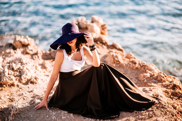 Fashionable girl in a big hat posing on a rock on the beach