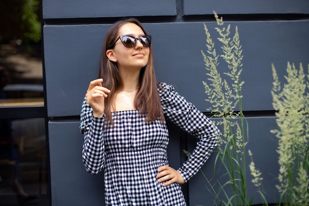 Fashionable girl in plaid dress and sunglasses looks into the sky and smiles on a black dark background