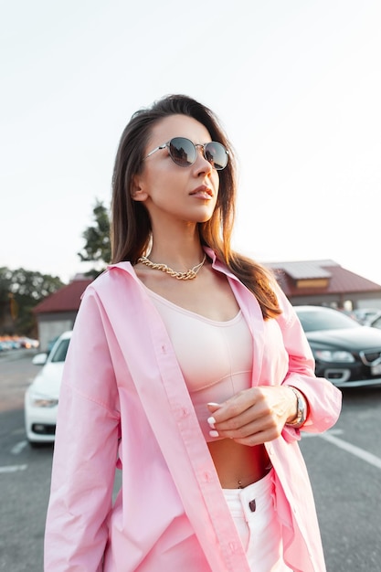 Fashionable glamorous beautiful woman with stylish sunglasses in trendy pink shirt and fashion top walks on the street Urban female style look clothes