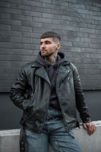 Fashionable handsome hipster man in stylish casual clothes with a leather jacket and hoodie sits near a black brick wall on the street