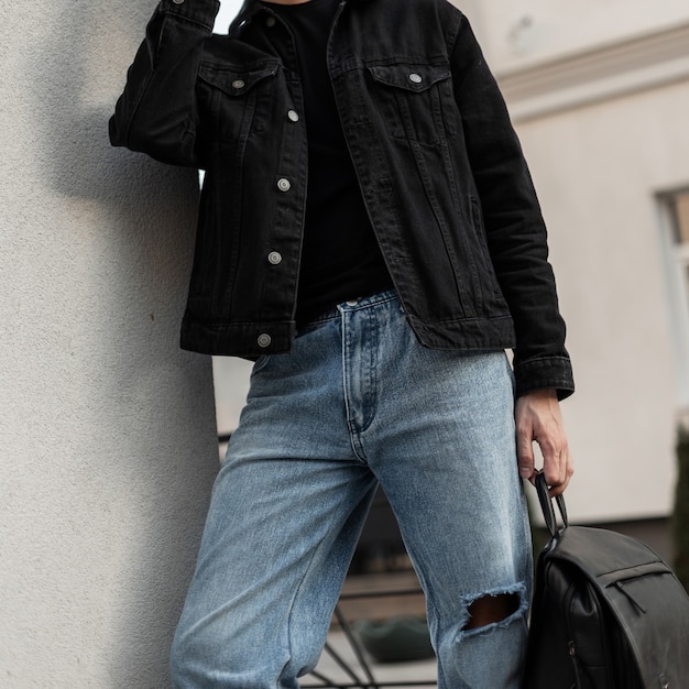 Fashionable handsome man in a black denim shirt with a black t-shirt and blue classic jeans holds a black leather backpack on the street