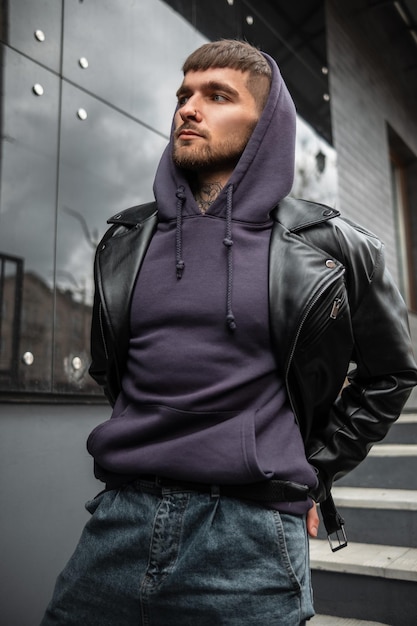 Fashionable handsome man model with black leather jacket hoodie and jeans walks near a black modern building in the city