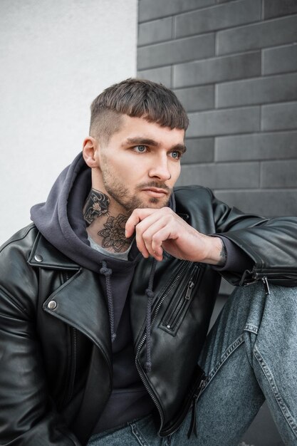 Fashionable handsome model man hipster with hairstyle and beard with a tattoo on his neck in a black leather jacket and hoodie sitting on the street