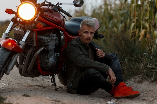 Fashionable handsome young man in military stylish khaki jacket with red sneakers sits and rests near a motorcycle with light outdoors