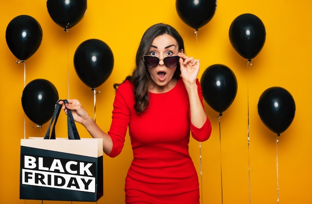 Fashionable happy gorgeous lady with a shopping bag in hand is posing in a good excited mood in the background of many helium balloons in black Friday