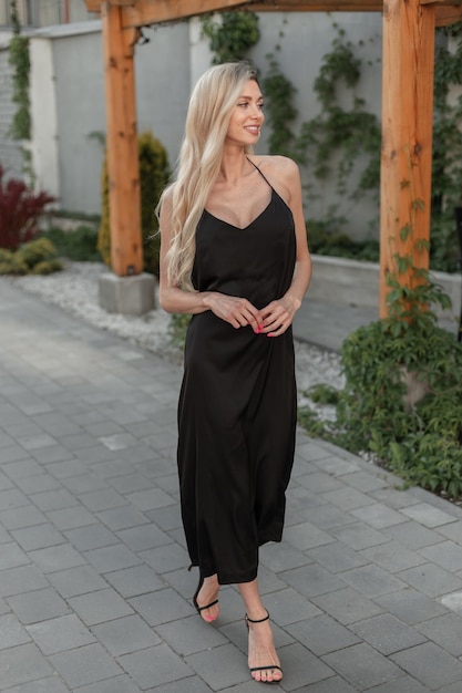 Fashionable happy pretty woman model with blonde hair in black stylish long strappy dress with shoes walks on the street