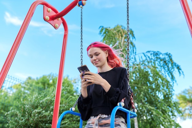 Fashionable happy teenage girl with dyed hair with smartphone riding swing typing text message communicating in social networks blogging on phone Teen lifestyle technology youth concept