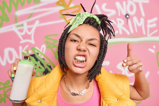 Fashionable hipster girl rapper with dreadlocks and golden teeth goes out to paint in public places holds spray aerosol creats graffiti on walls being passionate street artist