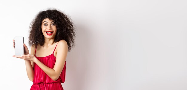 Photo fashionable lady in red dress and makeup showing mobile phone screen and smiling introduce smartphon