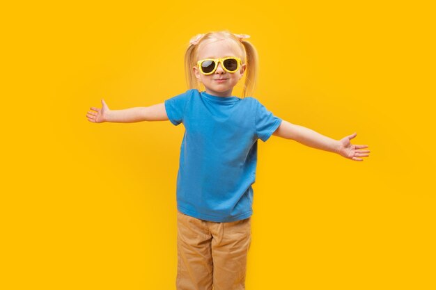 Fashionable little blonde girl in blue tshirt with sunglasses child raised hands up on yellow background and looking at camera