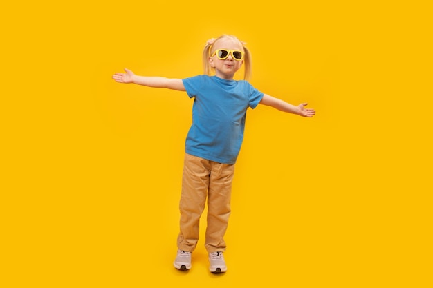 Fashionable little blonde girl in yellow pants and blue Tshirt with stylish sunglasses Child raised hands up on yellow background and looking at camera