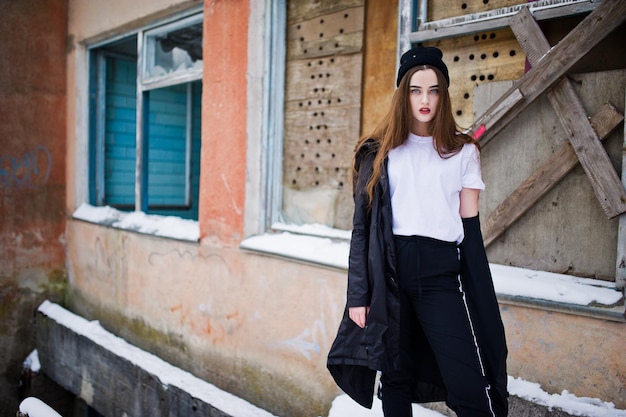 Fashionable long legs brunette model in long black cloak posed outdoor at winter day against old grunge wall with broken windows.