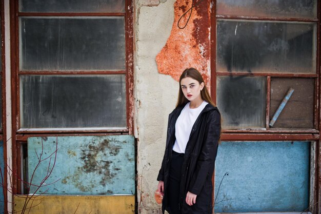 Fashionable long legs brunette model in long black cloak posed outdoor at winter day against old grunge wall with broken windows