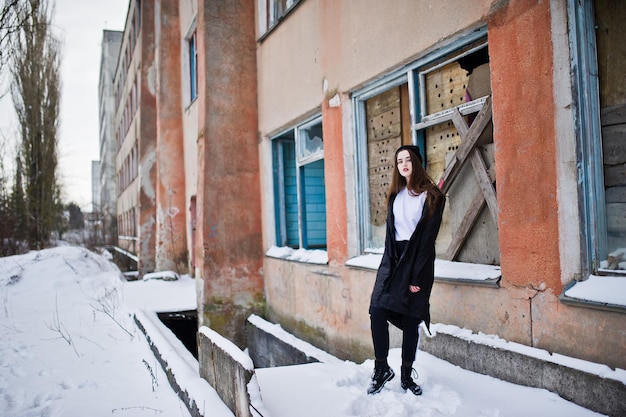 Modello bruna alla moda gambe lunghe in lungo mantello nero poste all'aperto al giorno d'inverno contro il vecchio muro di lerciume con finestre rotte