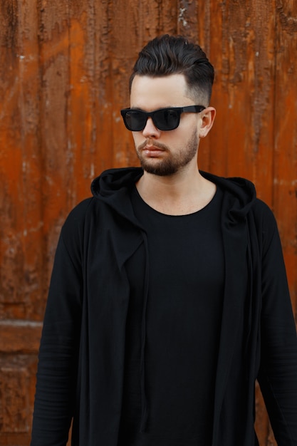 Fashionable man in sunglasses in a black hoody near a vintage wooden wall
