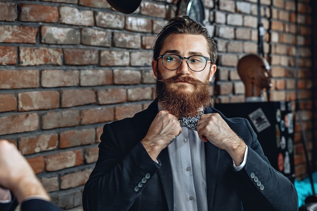 fashionable man with glasses, beard and mustache, looking at himself in the mirror and straightening a bow tie on his collar.