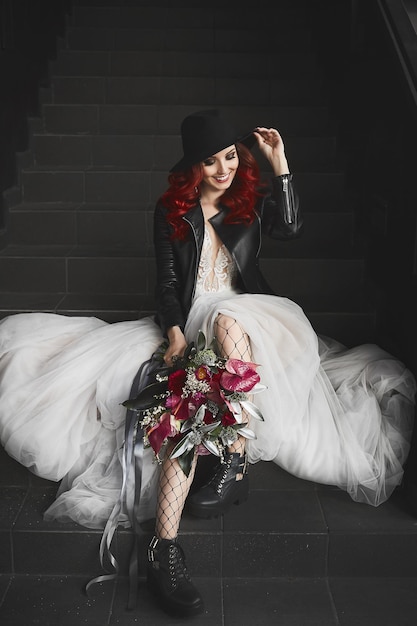 Fashionable model girl with red hair and beautiful smile in a white wedding dress and in a leather j...