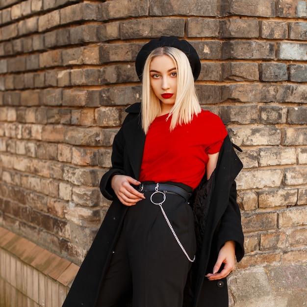 Fashionable modern young woman blonde with red lips in stylish clothes in retro style posing near a vintage brick building