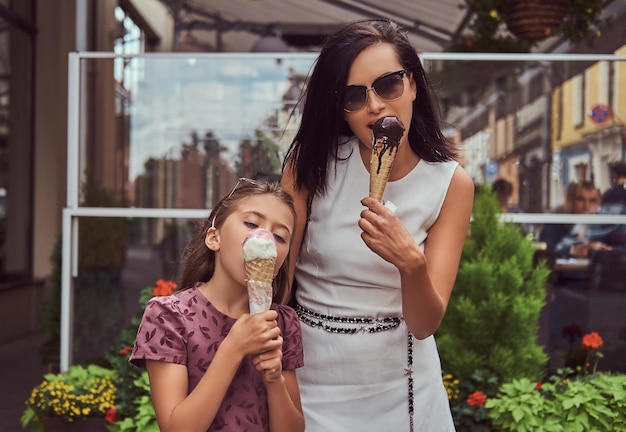 Fashionable mother and cute daughter enjoy ice cream on a hot summer day, standing near a cafe.