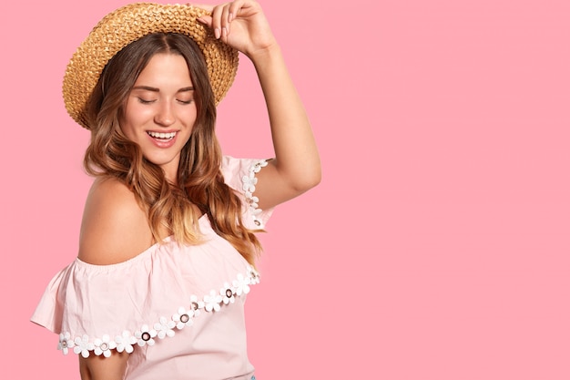 Fashionable pleased European woman keeps hand on straw hat