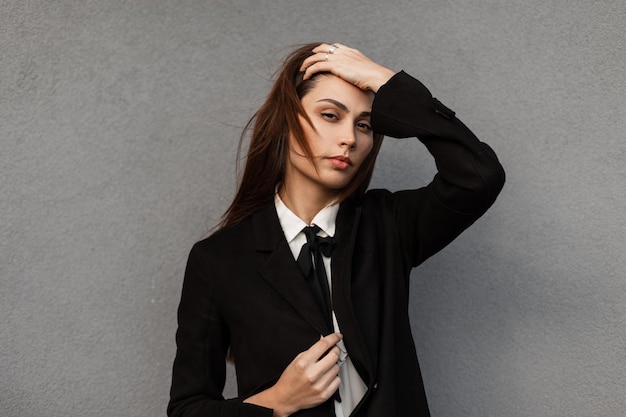 Модный портрет красивой молодой стильной деловой женщины в черном пальто и белой футболке с галстуком у серой стены в городе
