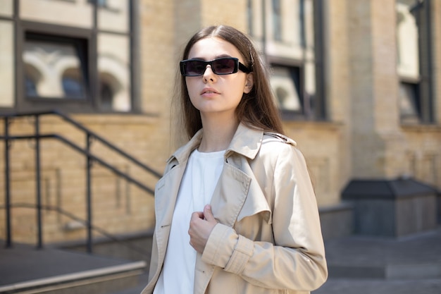 Fashionable portrait of a brunette girl in black sunglasses and a beige trench coat on a brick wall background, close-up