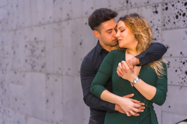 Fashionable pose of a Caucasian loving couple in a blue suit and green dress, hugging with closed eyes in the city