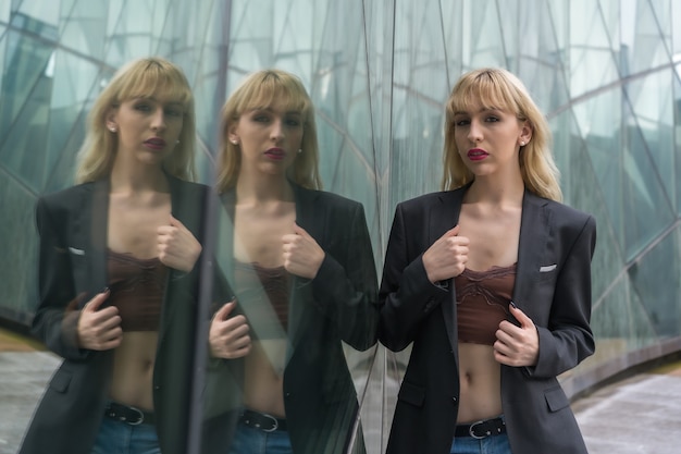 Fashionable pose of a young blonde in a glass buildings in the city, young man in a black suit looking at the camera reflected in the windows