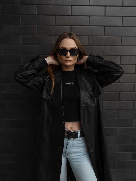 Fashionable pretty girl with black stylish sunglasses in black fashion long leather coat with tshirt and jeans stands near a black brick background Fashion style and beauty Dark clothes look