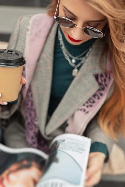 Fashionable redheaded girl in stylish clothes with a suit and scarf reading a magazine and drinking coffee