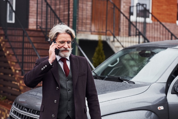 Fashionable senior man with gray hair and beard is outdoors on the street near his car have conversation by the phone.