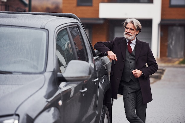 Fashionable senior man with gray hair and beard standing outdoors on the street near his car with keys in hand.