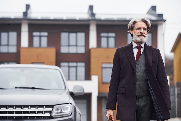 Fashionable senior man with gray hair and beard walking outdoors on the street near his car.