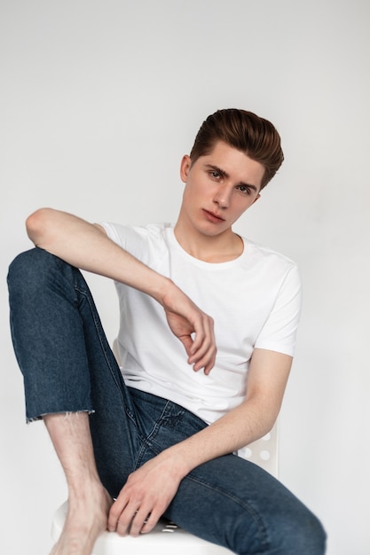 Fashionable serious young man in blue vintage jeans in stylish white t-shirt sits on chair on white chair in room. Studio portrait handsome guy in trendy clothes. New fashion collection casual wear.