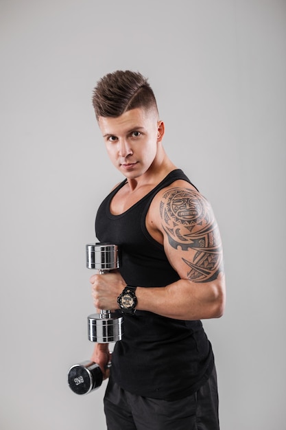 Fashionable stylish bodybuilder man model with hair in a black tank top with a muscular body is training with metal dumbbells on a gray background in the studio