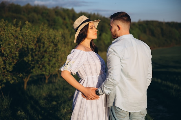 Fashionable and stylish happy pregnant woman and her husband dressed a pastel white and blue tone in the field on the sunset