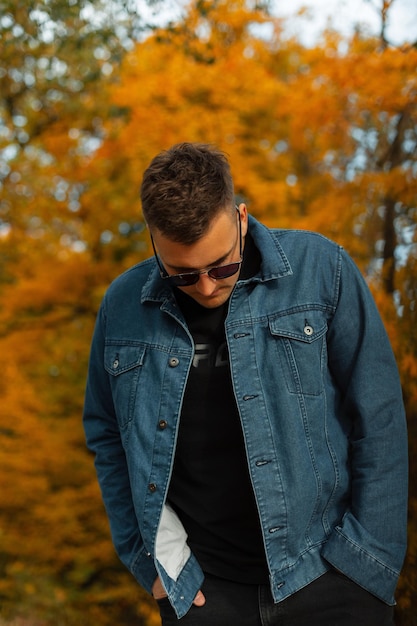 Uomo alla moda alla moda con capelli e occhiali da sole in abiti denim alla moda che cammina in un parco autunnale con fogliame luminoso