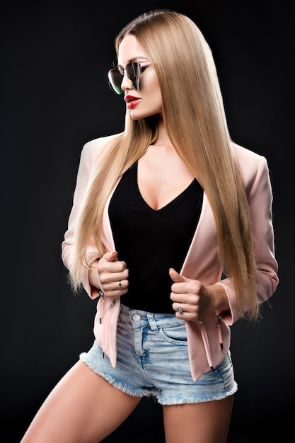 Fashionable stylish woman in denim outfit and a beautiful coat looking to the side at the mirrored sun glasses. The girl&#39;s red lips. Studio photo on a black background.