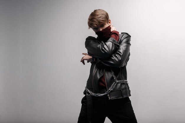 Fashionable stylish young man with a silver metal chain in a red vintage golf in a black leather stylish jacket in black stylish jeans posing against a white wall indoors. Modern fashion. Nice guy