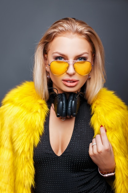 Модная стильная молодая женщина в наушниках с желтыми модными солнцезащитными очками и стильной желтой шубой на сером