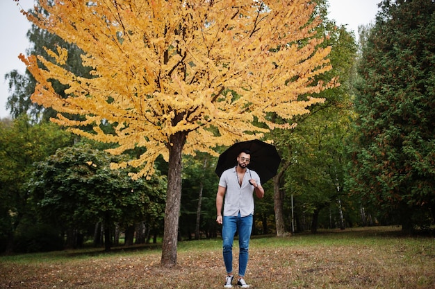 Модный высокий арабский бородатый мужчина в джинсах-рубашках и солнцезащитных очках позирует в осеннем парке с зонтиком на фоне желтых листьев дерева