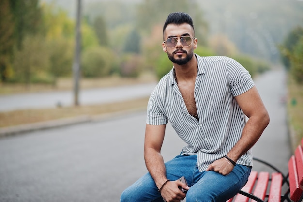 Fashionable tall arab beard man wear on shirt jeans and sunglasses posed on park at bench Hold his wallet at hand