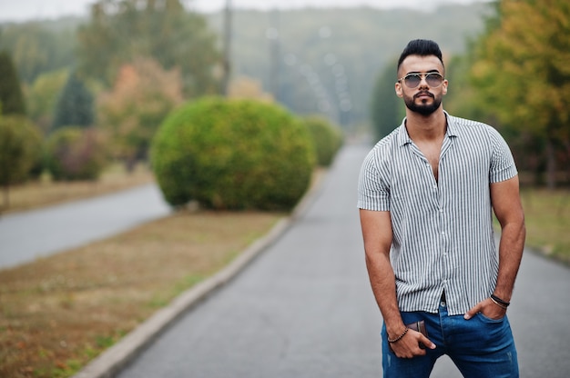 Moda uomo barbuto alto che indossa camicia, jeans e occhiali da sole poste sul parco e tenere il portafoglio a portata di mano